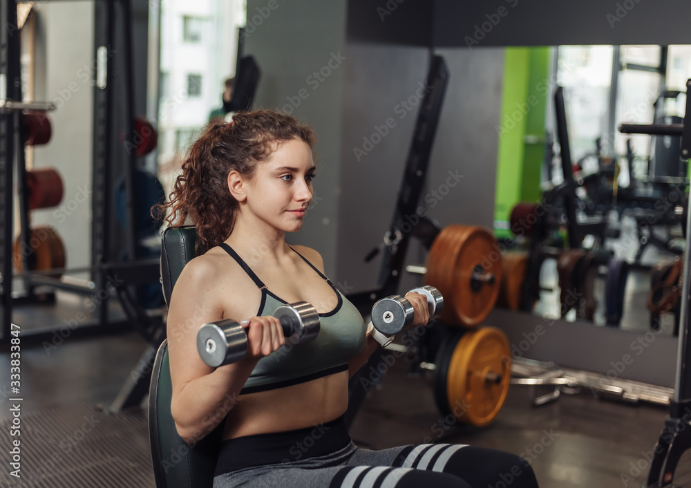 Fototapeta premium Young fit woman doing exercise lifting dumbbell with hands while sitting on a bench in the gym