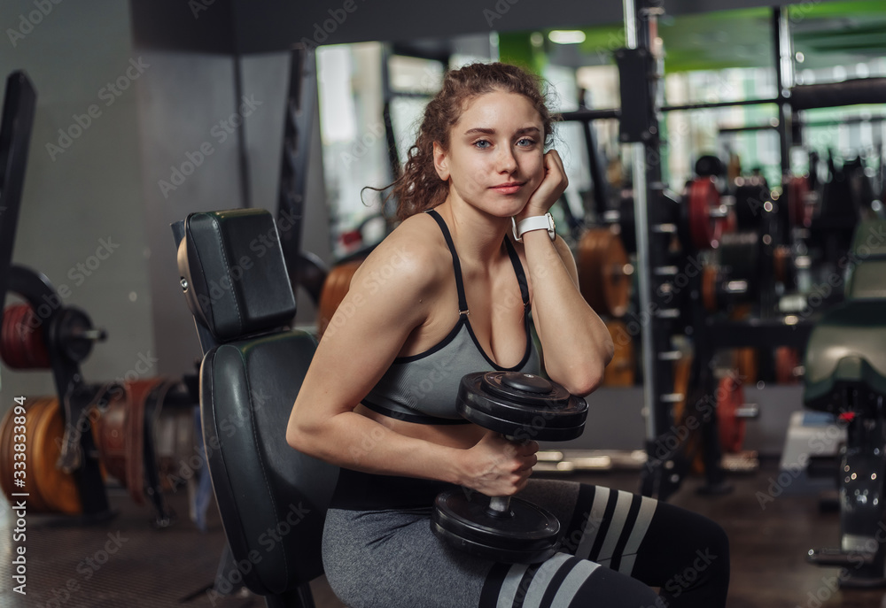 Fototapeta premium Tired young fit woman holding dumbbells and resting while sitting on a bench in the gym