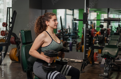 Cheerful young fit woman holding dumbbells while sitting on a bench in the gym