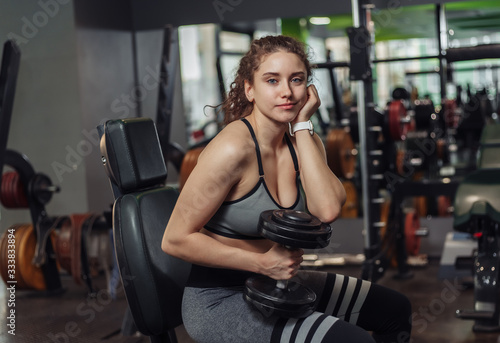 Tired young fit woman holding dumbbells and resting while sitting on a bench in the gym © splitov27