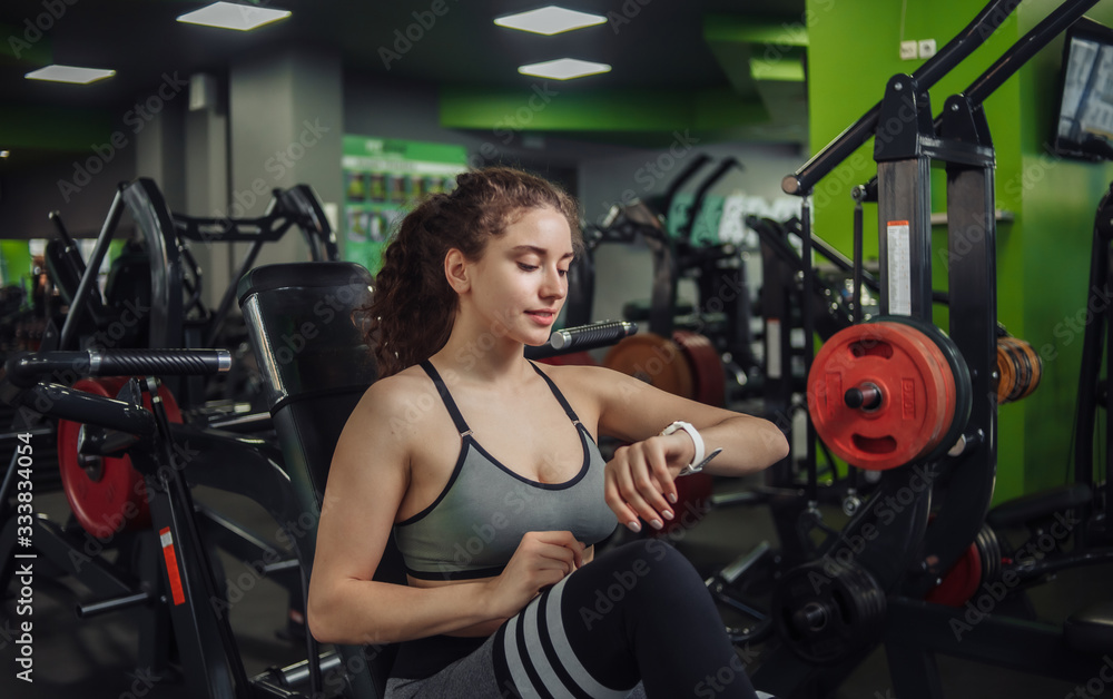 Young fit woman sitting on a bench in the gym and looks at the watch. Rest between exercises. Healthy lifestyle concept Young fit woman sitting on a bench in the gym and looks at the watch. Rest between exercises. Healthy lifestyle concept