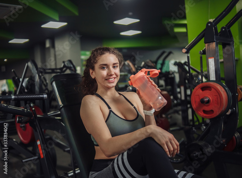 Young fit woman drinks water from a bottle while sitting on a bench in the gym. Rest between exercises. Healthy lifestyle concept