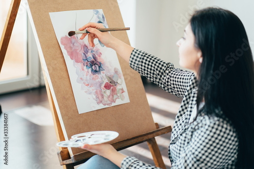 Artwork, creativity and inspiration. Young woman sitting at easel painting with watercolors. Drawing studio
