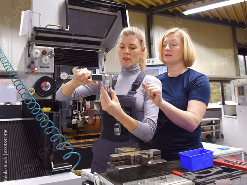 A young trainee receives tuition from her trainer. Both women work as computerized  numerical control engineers. They are seen at their high tech place of work.