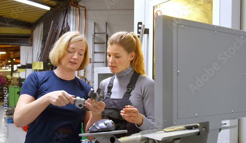 A young trainee receives tuition from her trainer. Both women work as computerized  numerical control engineers. They are seen at their high tech place of work.
