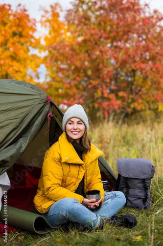 travel, trekking and hiking concept - young woman sitting near green tent and drinking tea in autumn forest