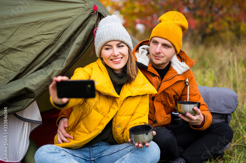 travel, trekking and hiking concept - portrait of couple sitting near green tent, drinking tea and taking selfie photo in autumn forest