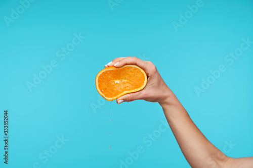 Beautiful fair-skinned pretty hand keeping her hand raised while holding fresh orange, squeezing it with fingers while isolated against blue background