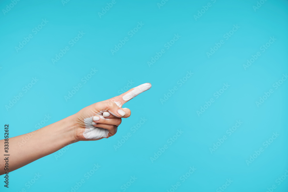 Close-up of young pretty lady's hand painted with white color keeping index finger raised while showing aside, posing against blue background Close-up of young pretty lady's hand painted with white color keeping index finger raised while showing aside, posing against blue background