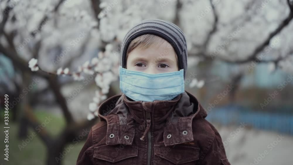 A 5-year-old child in a medical mask in a jacket and hat in the spring against the background of a blossoming tree. A 5-year-old child in a medical mask in a jacket and hat in the spring against the background of a blossoming tree.