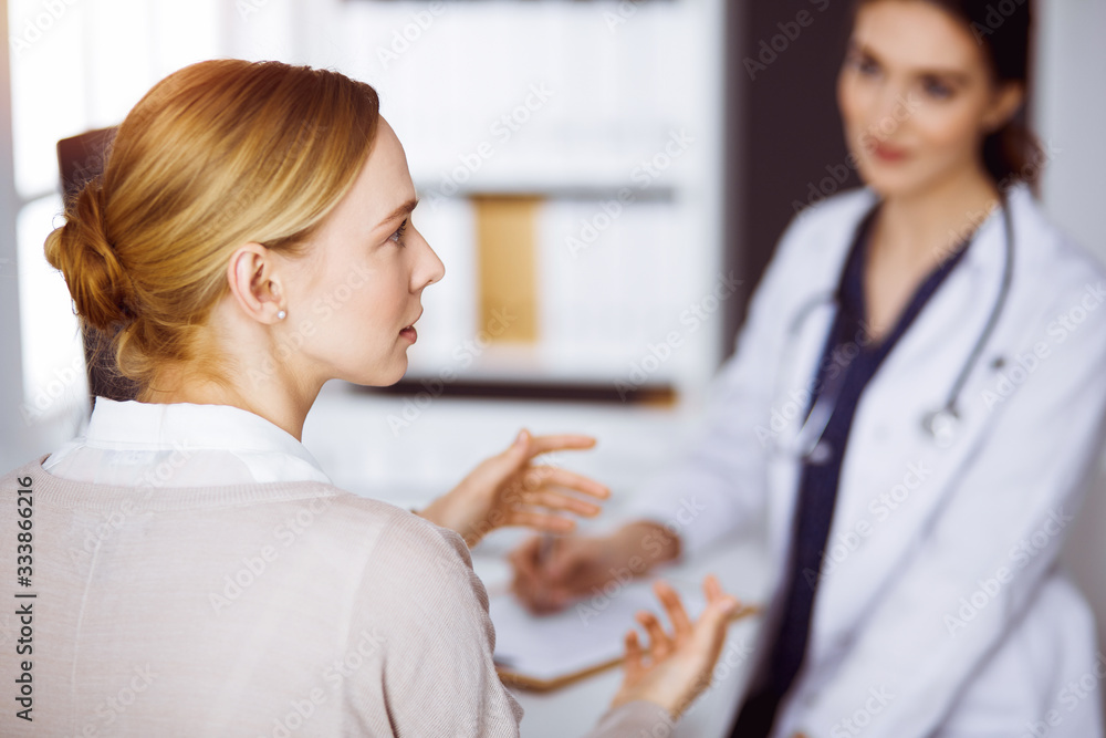 Obraz premium Cheerful smiling female doctor and patient woman discussing current health examination while sitting in sunny clinic. Medicine concept