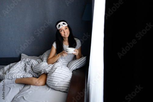 Happy beautiful woman in a sleep mask sitting on the bed in the house with a Cup of coffee/tea