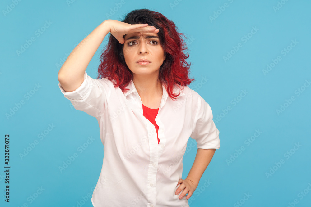 Distant future. Portrait of hipster woman with red hair holding hand over eyes and looking far away, expecting waiting something with worried expression. indoor studio shot isolated on blue background Distant future. Portrait of hipster woman with red hair holding hand over eyes and looking far away, expecting waiting something with worried expression. indoor studio shot isolated on blue background