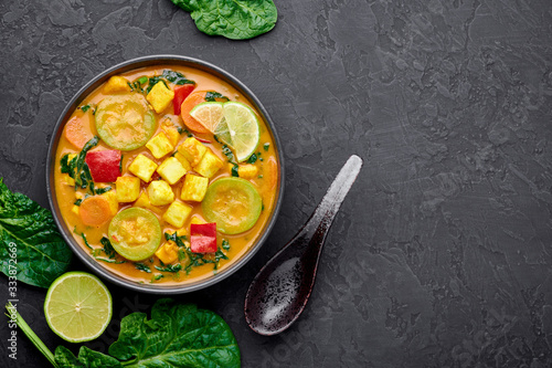 Yellow Veg Thai Curry with Tofu and vegetables in black bowl at dark slate background. Vegetarian Thai Curry with tofu, zucchini, pepper, spinach, carrot. Thai Food. Indian vegetable curry. Copy space