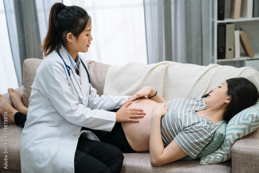 Side view of asian chinese female doctor examining expectant woman during visiting patient at home. elegant girl midwife putting hands on maternity abdomen of motherhood. future mom lying on couch. Side view of asian chinese female doctor examining expectant woman during visiting patient at home. elegant girl midwife putting hands on maternity abdomen of motherhood. future mom lying on couch.