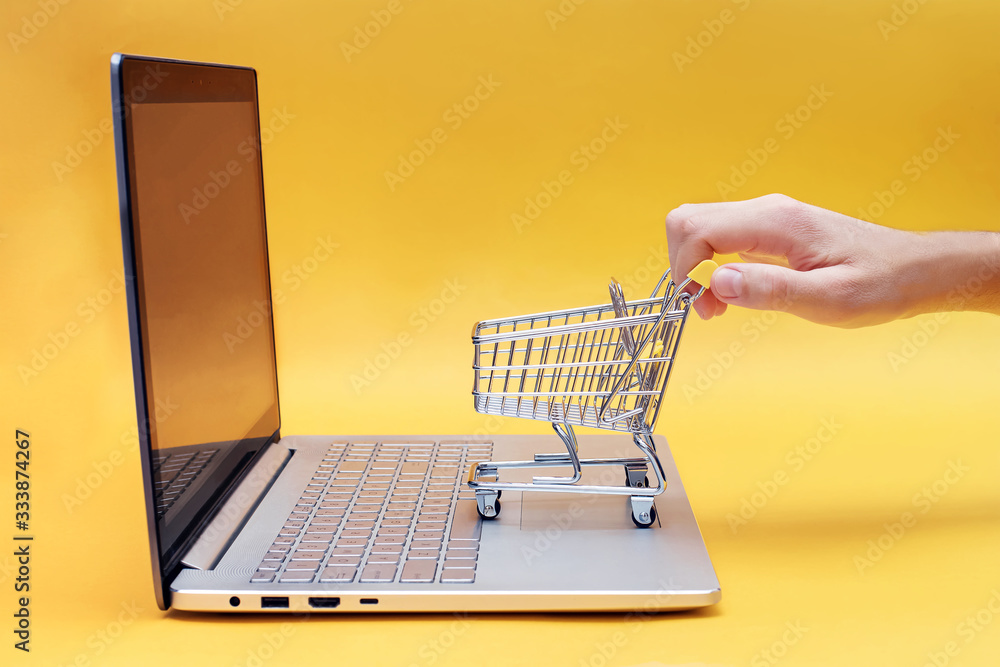 Hand holding mini shopping trolley near the opened laptop on yellow background.
