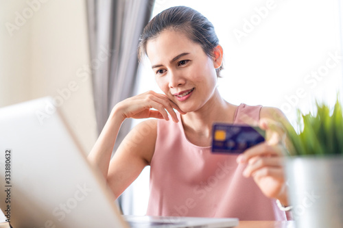 Asian young woman sit in chair, while using laptop for shopping online  from home. Girl hold credit card, smile and wait for delivery package box by shipping company. Shopaholic Lifestyle concept.