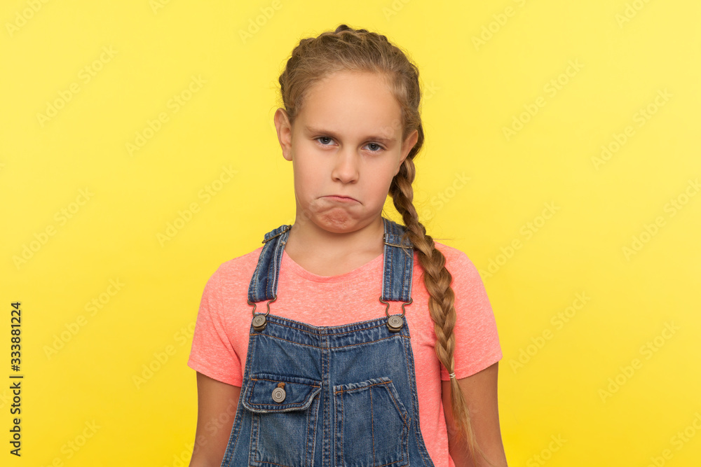Portrait of funny frustrated naughty little girl with braid in denim overalls looking at camera with comical resentful unhappy face expression. indoor studio shot isolated on yellow background Portrait of funny frustrated naughty little girl with braid in denim overalls looking at camera with comical resentful unhappy face expression. indoor studio shot isolated on yellow background