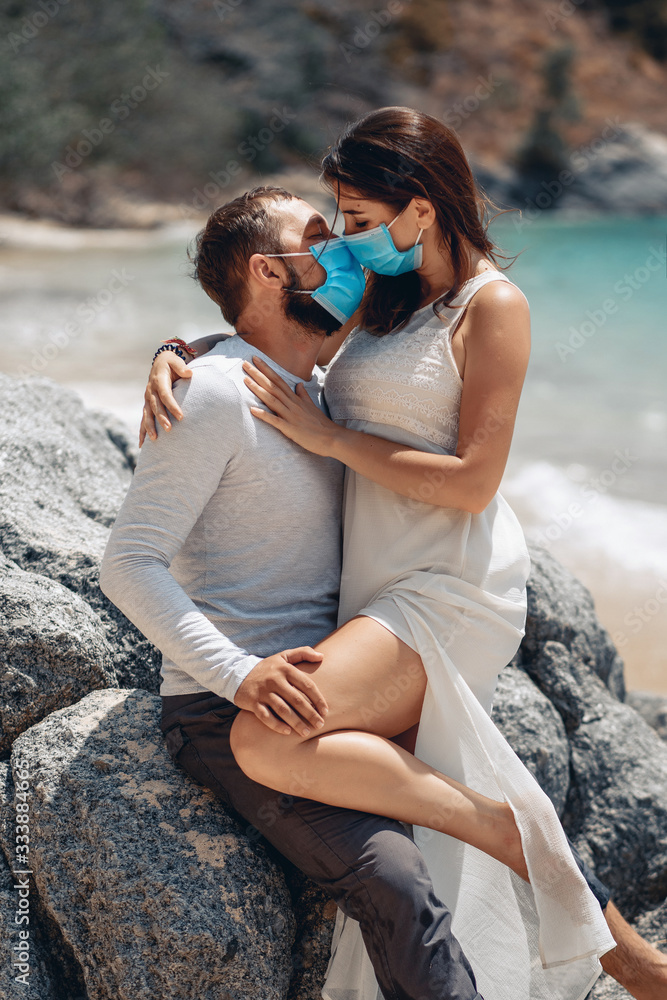 Portrait of a beautiful couple kissing sexually on the beach next to the azure sea while wearing a face mask that protects them from infection because of the outbreak of coronovirus infection COVID-19 Portrait of a beautiful couple kissing sexually on the beach next to the azure sea while wearing a face mask that protects them from infection because of the outbreak of coronovirus infection COVID-19