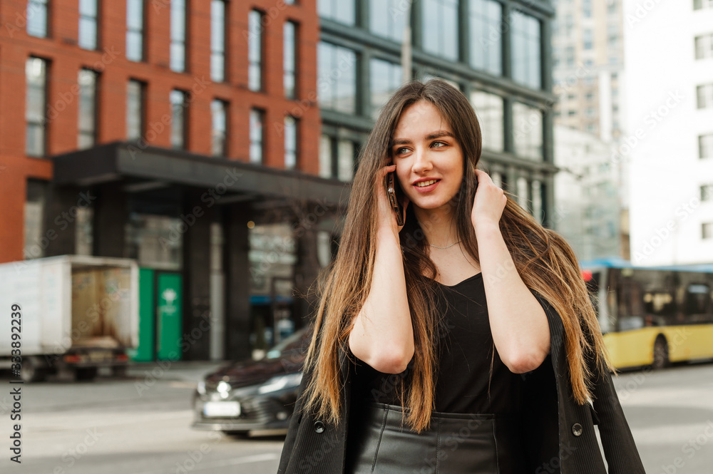 Beautiful girl with long hair is calling on the background of the street landscape, looking away with a smile on her face. Beautiful businesswoman in dark casual clothes talking on phone.Copy space
