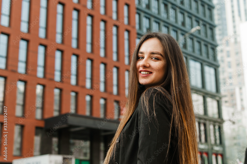 Positive girl in formal dress looks in camera and smiles,background of the metropolitan architecture.Street portrait happy young business woman in a dark jacket on the background of a modern building