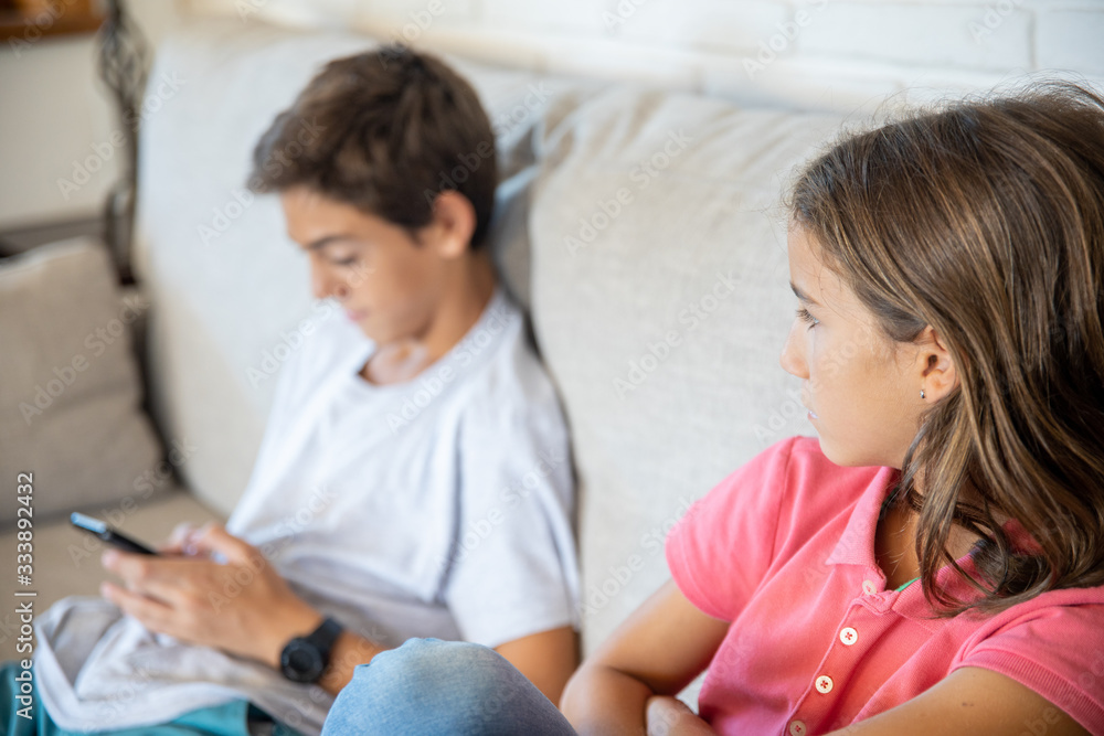 Happy boy playing with smartphone next to his angry sister looking at him Happy boy playing with smartphone next to his angry sister looking at him