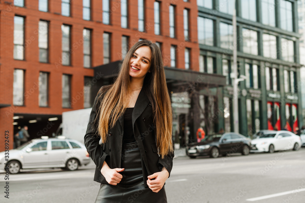 Street portrait of an attractive young business woman in formal wear, wearing a black jacket and skirt, looking in camera with a smile on face.Background of cityscape. Business portrait beautiful lady