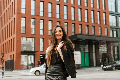 Portrait of a happy model in formal clothes walking down the metropolis street with a smile on her face and looking away. Positive young businesswoman, street portrait on cityscape background.