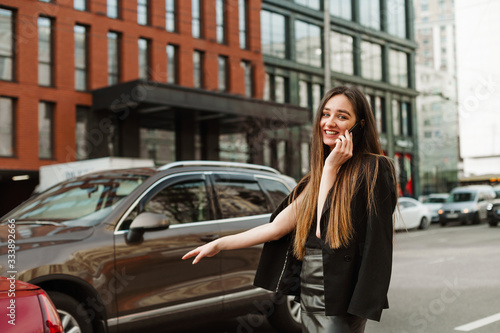 Positive girl in formal clothes stands on the road and catches a taxi with her hand raised. Cheerful young businesswoman in dark clothing is calling on the phone and hand gesture stops a taxi.