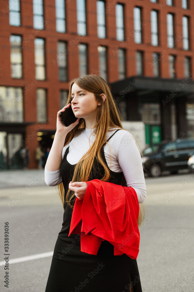 Portrait of a concentrated lady in casual clothes standing on the street with a jacket in her hand and calling on the background of a street landscape. Student girl talking on the phone on the street.
