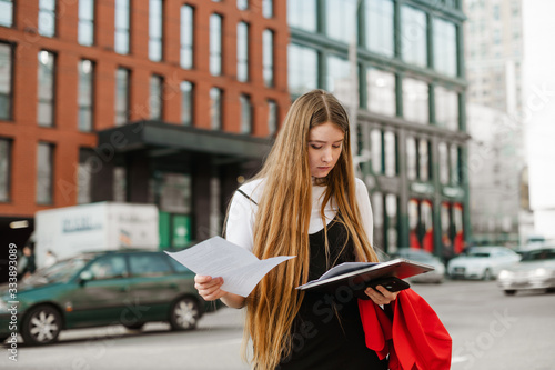 Busy young girl in casual clothes stands on the street of a metropolis with a laptop and papers in her hands,reads information from papers.Student studies on the way to university
