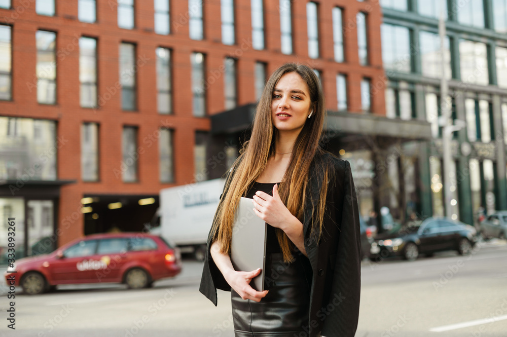 Pretty young businesswoman in dark formal wear posing with a laptop in her hands on the city street, looking into the camera with a smile on her face, wearing wireless headphones. Business people. Pretty young businesswoman in dark formal wear posing with a laptop in her hands on the city street, looking into the camera with a smile on her face, wearing wireless headphones. Business people.