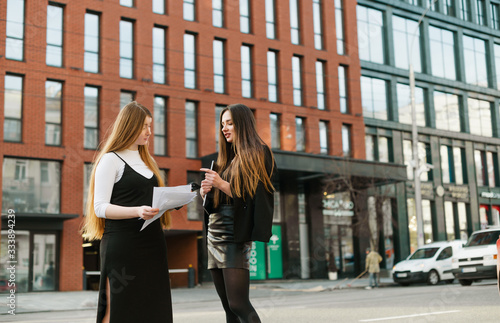 Two businesswomen stand on street with a laptop and business papers in their hands and talk.Girlfriends office workers discussing work on a background of urban landscape.Business meeting on the street