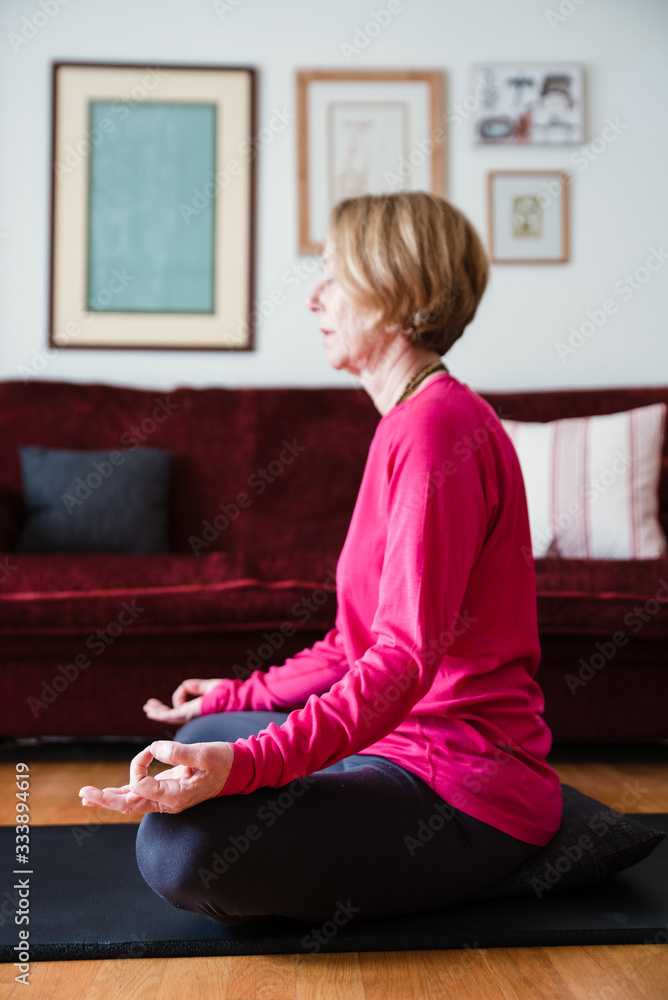 Middle aged woman sitting in lotus position on yoga mat in her living room. Middle aged woman sitting in lotus position on yoga mat in her living room.
