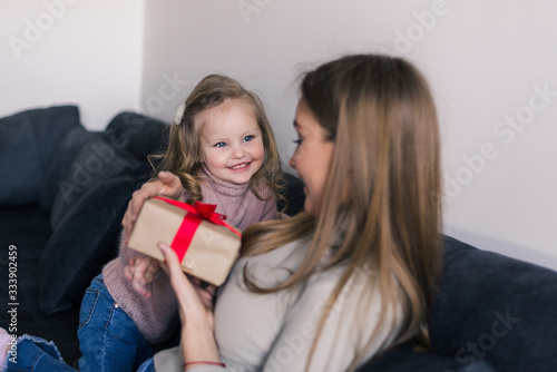 Happy young girl surprising her mother with gift at home in living room. Mother day.