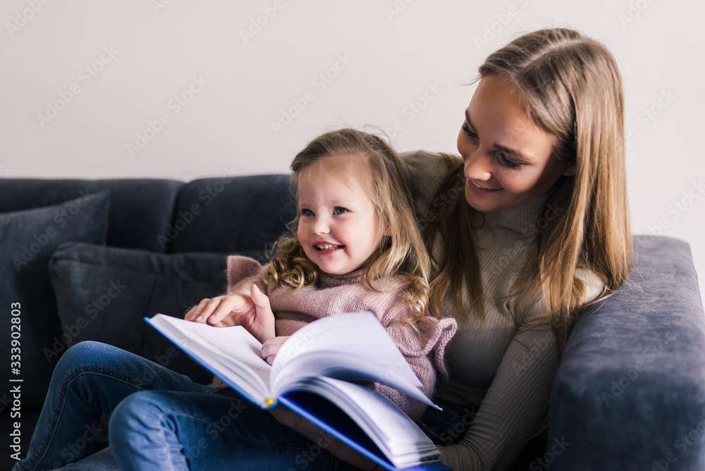 Happy mother and little daughter reading book, sitting on comfortable couch in living room. Smiling mum teaching little girl child, family spending weekend at home together. Children education