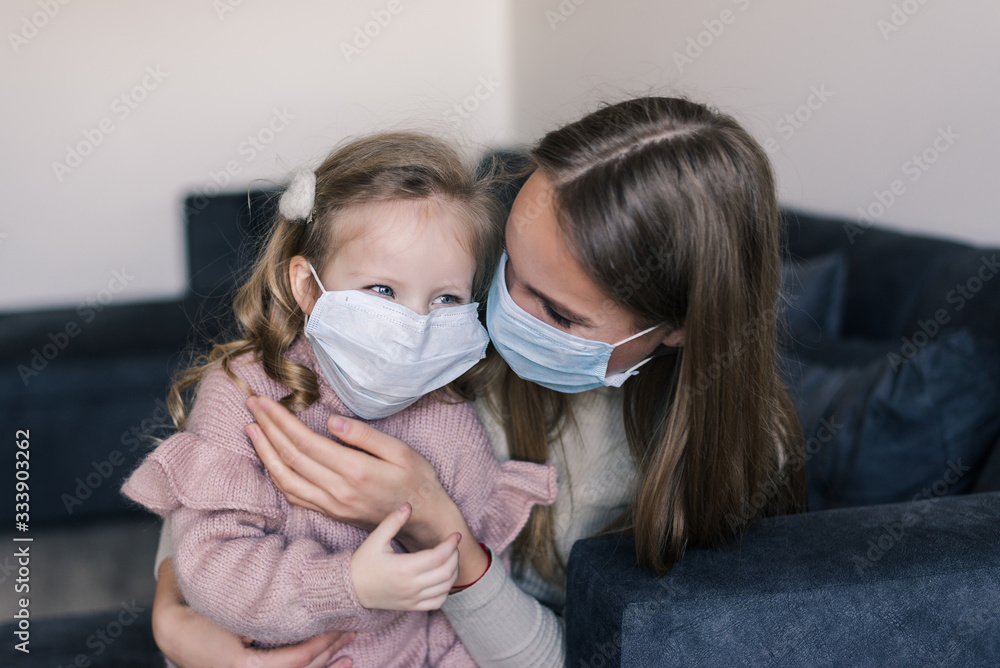 Cute little girl and mother wearing face mask, sitting on bed at home, consoling sad preschool daughter. Concept of coronavirus or COVID-19 pandemic disease symptoms. Self isolation, home quarantine.