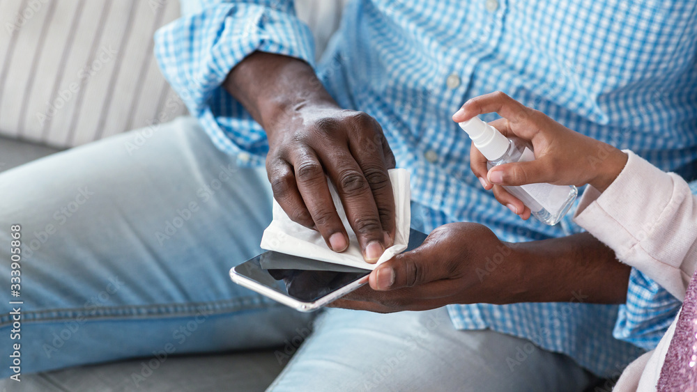 Infection prevention. African American man and little child cleaning mobile phone with sanitizer, closeup. Empty space