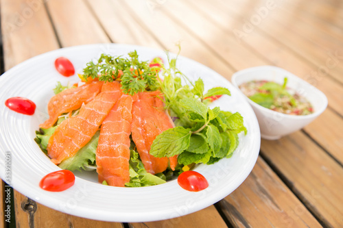 raw fresh salmon salad top with parsley decorated by mint and tomato in a white plate on wood table serve with thai style sause 