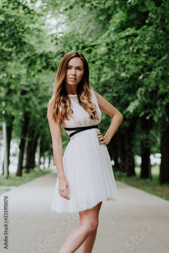 Outdoor fashion portrait of young beautiful happy smiling lady wearing white dress, stylish hairstyle, posing in park. City