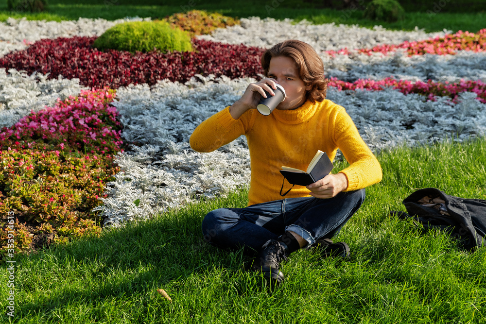 The young man drinks coffee and reads the book in the park, he smiles, has a rest, is dressed in a yellow sweater, flowers and grass on background, sunny day The young man drinks coffee and reads the book in the park, he smiles, has a rest, is dressed in a yellow sweater, flowers and grass on background, sunny day