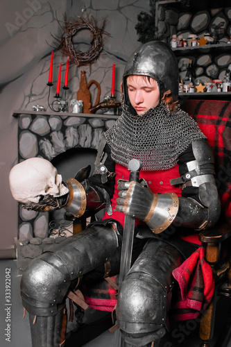 Young knight in armor 14th century with a sword in one hand and a skull in the other hand, sitting in a rocking chair by the fireplace. photo