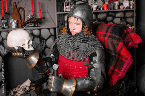 Young knight in armor 14th century with a sword in one hand and a skull in the other hand, sitting in a rocking chair by the fireplace. photo