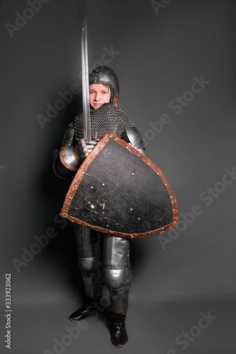 Photo A young knight in medieval armor armed with a sword and shield standing on a gray background