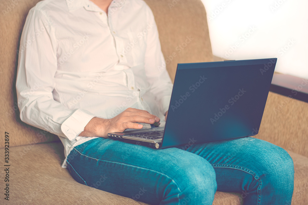 Fototapeta premium young man in white shirt and blue jeans working on laptop, writes in notebook in home office sitting on cozy beige sofa Stay at home, online work, distance learning concept