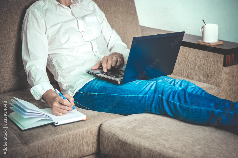 Naklejka premium young man in white shirt and blue jeans working on laptop, writes in notebook in home office sitting on cozy beige sofa Stay at home, online work, distance learning concept