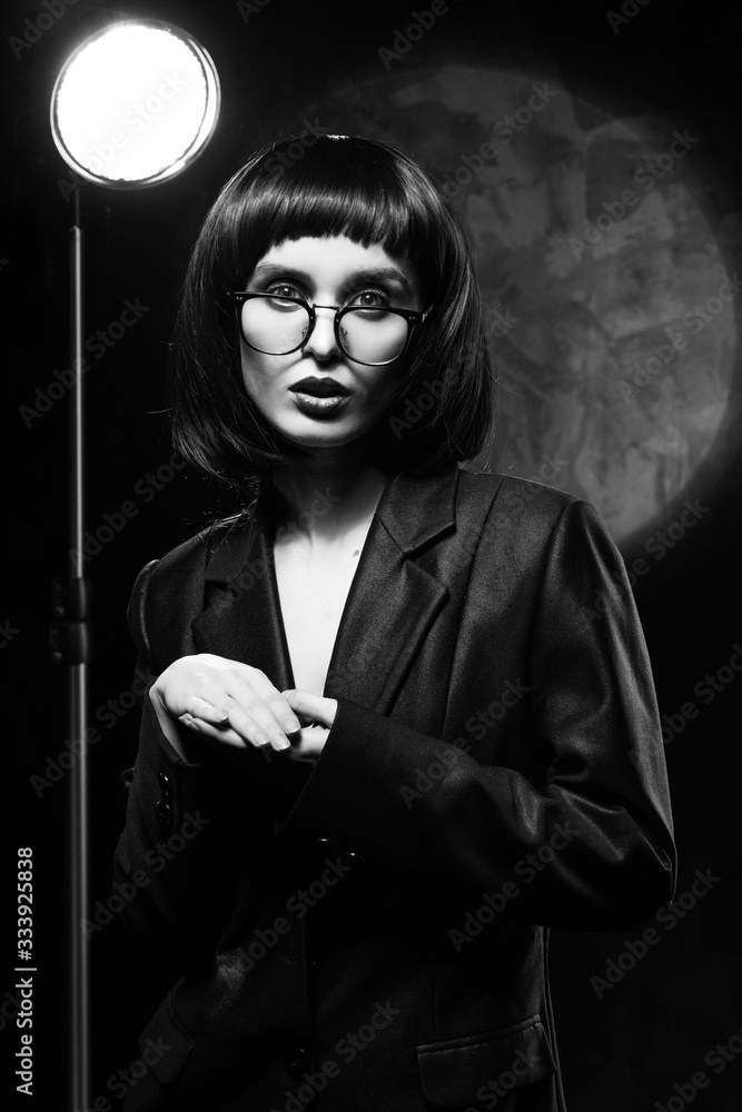 Beautiful braless slim girl with red lips, wearing a unbuttoned black blazer, glasses and brunette wig on a dark background, posing next to the light lamp. Monochrome, trendy, artistic design.