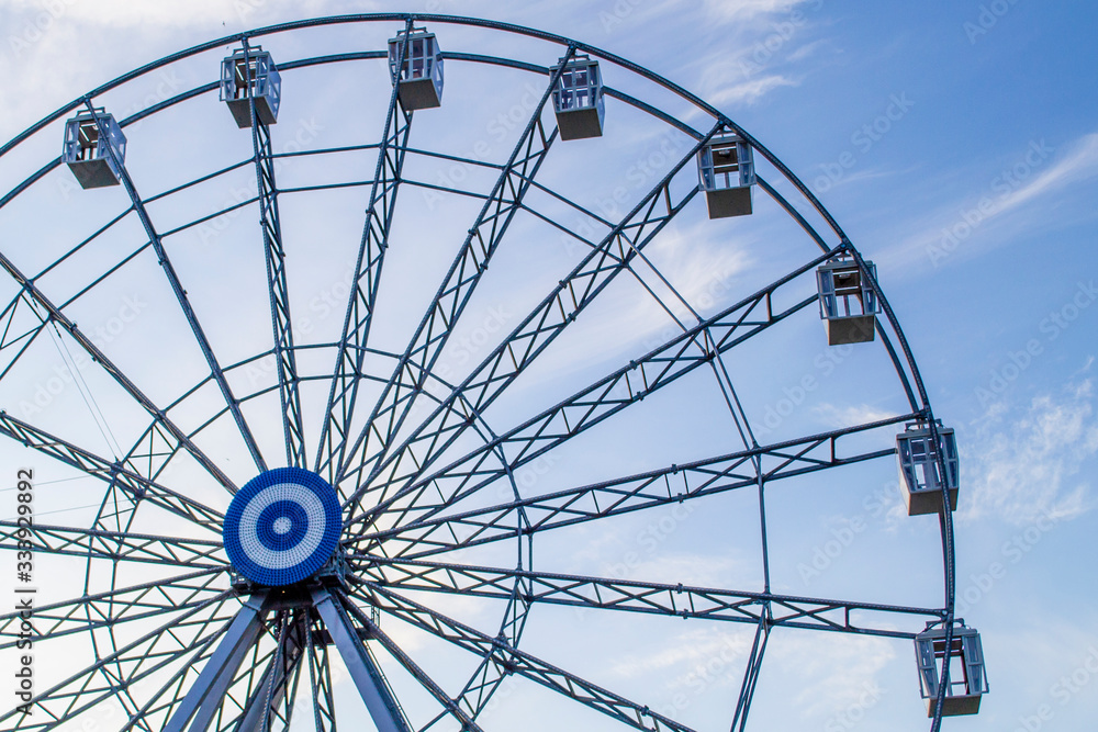 Fototapeta premium Ferris wheel on a background of blue sky and white clouds in the evening. Amusement park in the park Youth, in Kaliningrad, Russia. The concept of relaxation and fun.