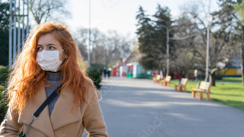 Fotografie Beautiful red haired girl wearing surgery mask walking on empty streets due to epidemic in city