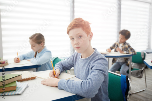 Portrait of modern red-haired schoolboy sitting at desk in classroom and looking at camera while taking notes, copy space